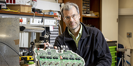 A man smiles at the camera. In front of him is a large electronic component.