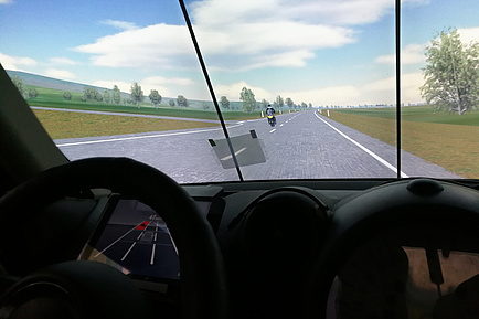 Cockpitperspektive, hinter der Windschutzscheibe ist eine virtuelle Landschaft, auf der Freilandstraße fährt ein gelbes Motorrad, das das Auto offensichtlich gerade überholte. Am Instrumentenbrett hinter dem Lenkrad wird die Straße und das Motorrad als roter Punkt angezeigt. 
