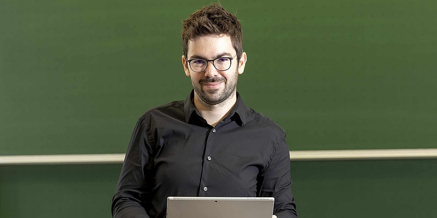 Ein Mann mit dunklen Haaren, dunkler Brille und Vollbart blickt in die Kamera. Er hält ein Notebook in den Händen. Im Hintergrund sieht man eine grüne Tafel. 