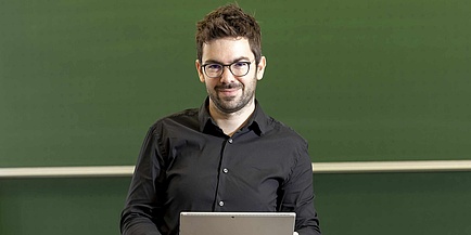 Ein Mann mit dunklen Haaren, dunkler Brille und Vollbart blickt in die Kamera. Er hält ein Notebook in den Händen. Im Hintergrund sieht man eine grüne Tafel. 