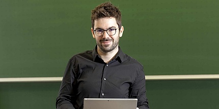 Ein Mann mit dunklen Haaren, dunkler Brille und Vollbart blickt in die Kamera. Er hält ein Notebook in den Händen. Im Hintergrund sieht man eine grüne Tafel. 
