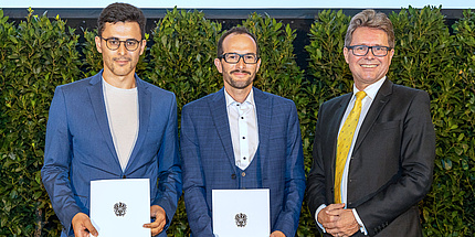 Two men in blue suits stand next to a third man, each holding a certificate.