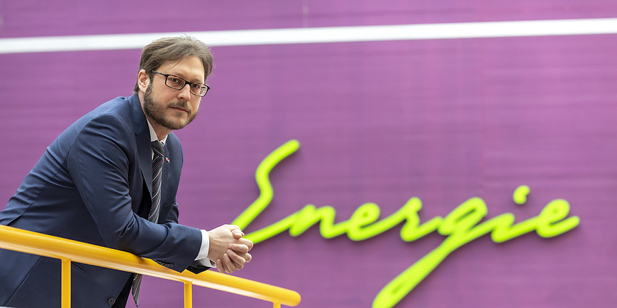 Ein Mann lehnt an einem Geländer und schaut in die Kamera, im Hintergrund steht das Wort Energie an der Wand geschrieben.
