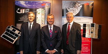 Three men in suits stand in front of two roll-ups of the TU Graz and are flanked by two satellite models.