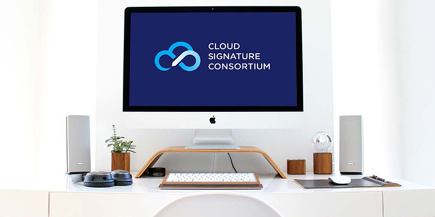 das Bild zeigt einen Computerarbeitsplatz mit einem Bildschirm und einer Tastatur sowie zahlreichen Büroutensilien. Am Bildschirm ist das Logo des Cloud Signature Consortiums zu sehen.