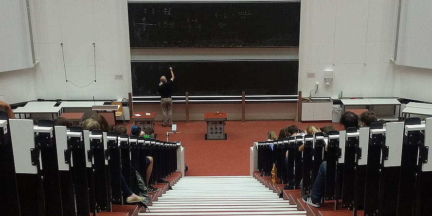 Auditorium. White stairs lead to the blackboard. A man is writing white letters on the blackboard. Students are sitting on the left and right of the stairs.