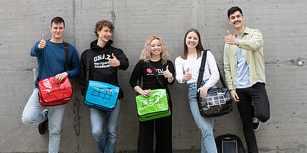 Zwei Studentinnen und drei Studenten mit bunten TU Graz-Planentaschen gut gelaunt vor einer Betonwand.