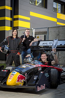 Man with a trophy in the cockpit of a racing car, next to him a woman in a racing suit and a man with tools.