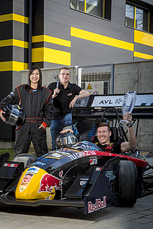 Mann mit Pokal im Cockpit eines Boliden, daneben eine Frau im Rennanzug und ein Mann mit Werkzeug.