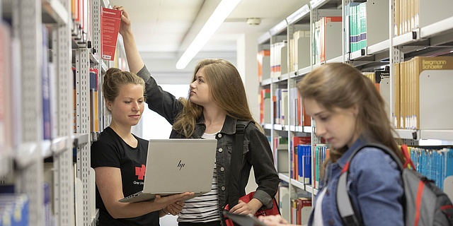 Drei Studierende stehen zwischen Bücherregalen, eine Person zieht ein Buch aus dem Regal, eine Person hält einen Laptop, die andere Person hält ein Tablet.