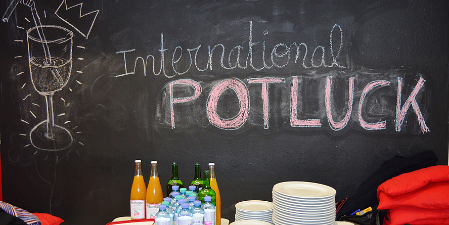 Angerichtetes Buffet mit Getränken und leeren Tellern vor einer Tafel mit der Aufschrift „International Potluck“.
