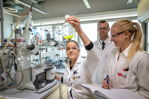 Lady with laboratory glass flask, another one taking notes, behind her the head of research