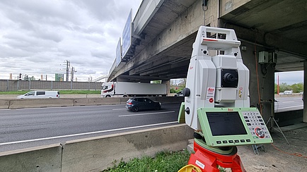 Ein Vermessungsgerät steht vor einer Autobahnbrücke.