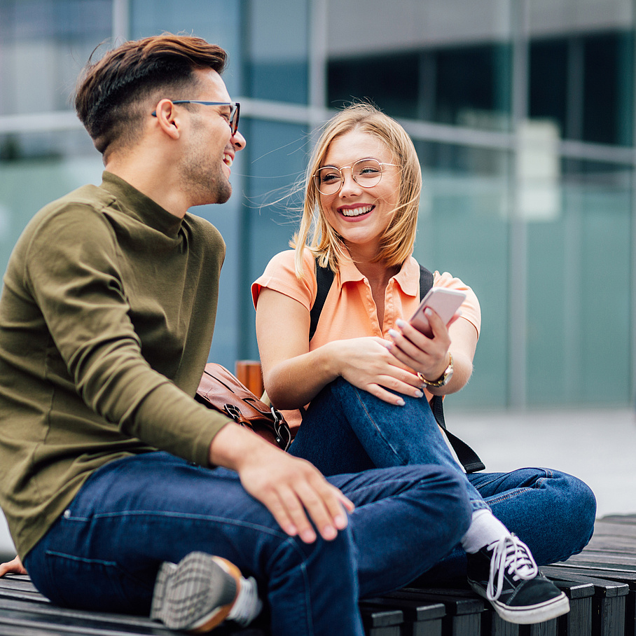 Two teenagers sit on a park bench and look at each other.