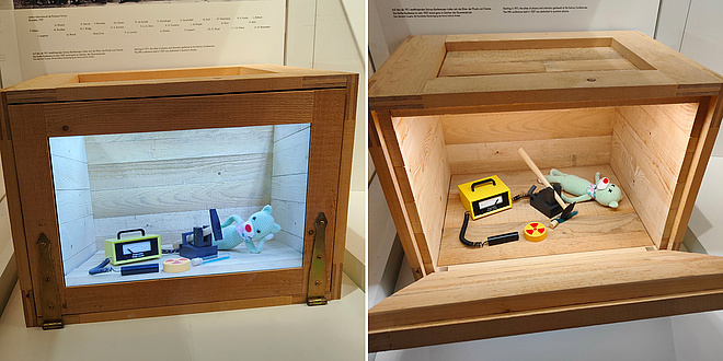 Zwei nebeneinander montierte Bilder. Links ist ein geschlossener Holzkasten mit einem Glasfenster zu sehen, in dem Kasten sieht man eine entspannt liegende Katzenpuppe mit einem Geigerzähler sowie einem Hammer, der über einer Giftkanüle schwebt. Rechts ist der geöffnete Holzkasten zu sehen, allerdings hat darin der Hammer die Giftkanüle zerstört und die Katzenpuppe wird als verstorben dargestellt.