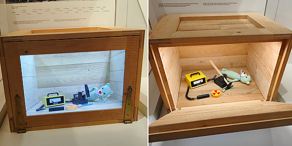 Zwei nebeneinander montierte Bilder. Links ist ein geschlossener Holzkasten mit einem Glasfenster zu sehen, in dem Kasten sieht man eine entspannt liegende Katzenpuppe mit einem Geigerzähler sowie einem Hammer, der über einer Giftkanüle schwebt. Rechts ist der geöffnete Holzkasten zu sehen, allerdings hat darin der Hammer die Giftkanüle zerstört und die Katzenpuppe wird als verstorben dargestellt.