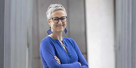 Eine Frau mit dunkler Brille, und kurzen grauen Haaren hält ihre Arme verschränkt. Sie lehnt an einer Wand und blickt in die Kamera. Die Frau trägt ein blaues Shirt und blaue Ohrringe.