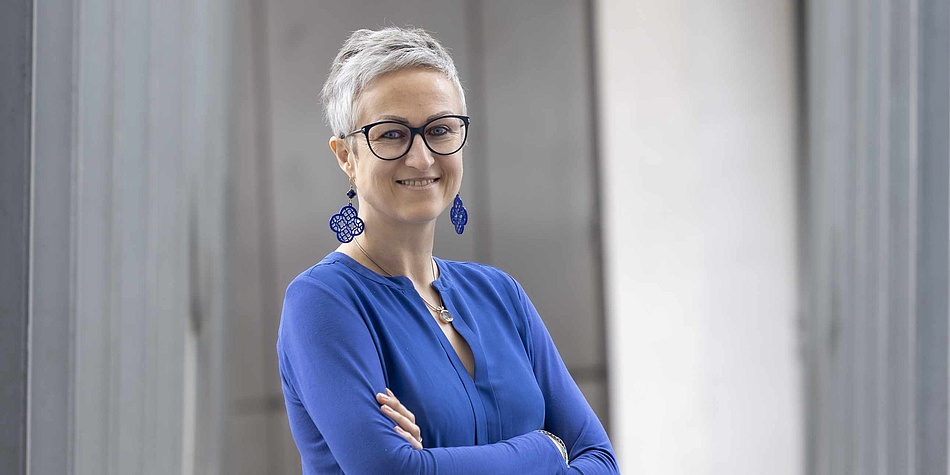 Eine Frau mit dunkler Brille, und kurzen grauen Haaren hält ihre Arme verschränkt. Sie lehnt an einer Wand und blickt in die Kamera. Die Frau trägt ein blaues Shirt und blaue Ohrringe.