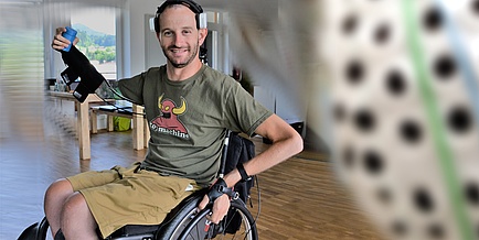 A young man is sitting in a wheelchair, holding his right arm in the air to hold a can.