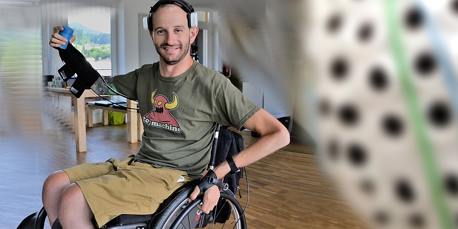 A young man is sitting in a wheelchair, holding his right arm in the air to hold a can.