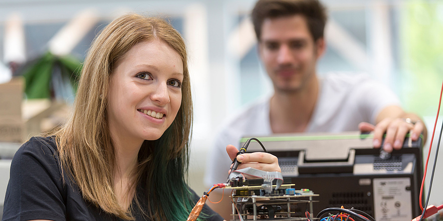 A young woman is working on an electric device and smilingly looks into the camera. In the background a young man is watching her.