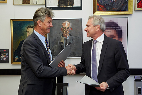 Two gentlemen in suits look at each other and shake hands. Both are holding a document folder in their other hand.