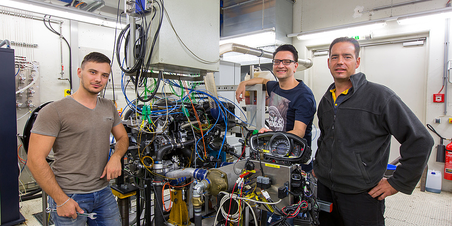 Three men in front of a device with many colourful cables, on the left in a brown T-shirt, in the middle a gentleman with glasses and on the right a gentleman with a grey jacket