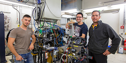 Three men in front of a device with many colourful cables, on the left in a brown T-shirt, in the middle a gentleman with glasses and on the right a gentleman with a grey jacket