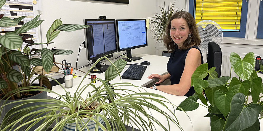 Eine Frau sitzt an einem Schreibtisch vor ihrem Computer. Es sind viele grüne Pflanzen aufgestellt. 