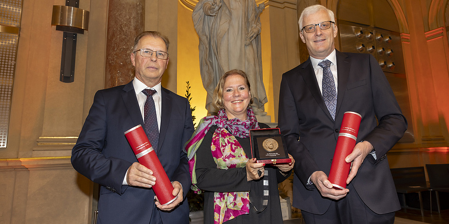 Zwei Männer und eine Frau lächeln in die Kamera. Die Männer halten Rollen mit Urkunden, die Frau eine Medaille