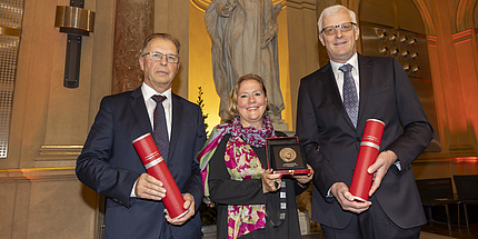 Zwei Männer und eine Frau lächeln in die Kamera. Die Männer halten Rollen mit Urkunden, die Frau eine Medaille