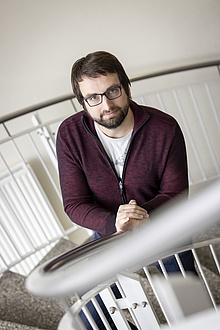 A man with a beard and glasses stands on a staircase and looks into the camera.