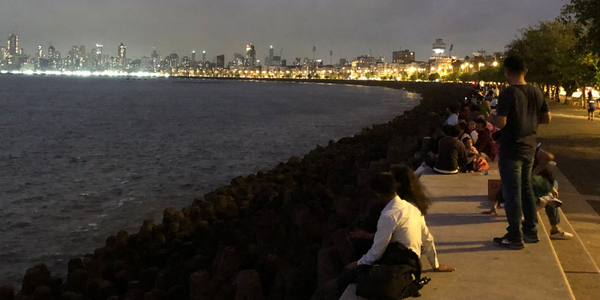 People are sitting at the edge of the harbour, looking out towards the open sea; in the background, you can see the brightly lit city at night.