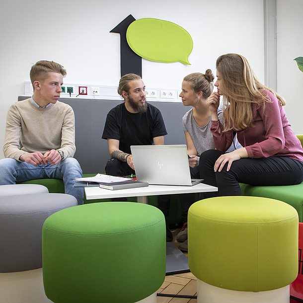 Two young men and two young women sit on green stools and a grey sofa. They talk to each other. On the wall above them is a green speech bubble. On a small table in front of them there is an open laptop.