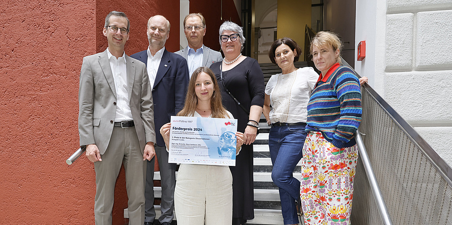 7 Personen blicken in die Kamera. Die Gruppe steht auf einer Stiege, in der Mitte steht eine junge Frau mit langen dunklen Haaren. Sie hält eine Urkunde mit dem Titel „Förderpreis 2024“ in der Hand. 