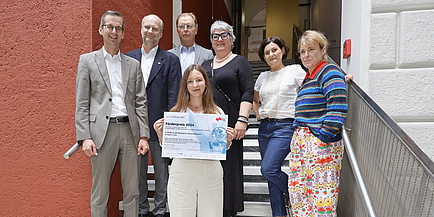 7 Personen blicken in die Kamera. Die Gruppe steht auf einer Stiege, in der Mitte steht eine junge Frau mit langen dunklen Haaren. Sie hält eine Urkunde mit dem Titel „Förderpreis 2024“ in der Hand. 