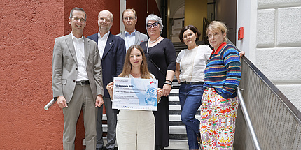 7 Personen blicken in die Kamera. Die Gruppe steht auf einer Stiege, in der Mitte steht eine junge Frau mit langen dunklen Haaren. Sie hält eine Urkunde mit dem Titel „Förderpreis 2024“ in der Hand. 