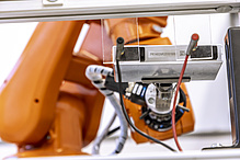 Ein Roboterarm hält eine flache Batterie fest, an der zwei Kabel angeschlossen sind.