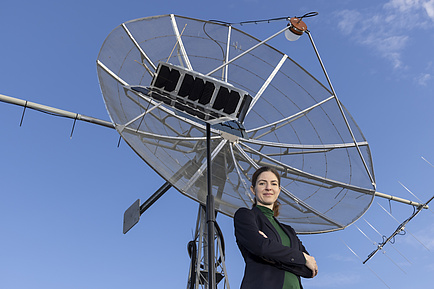 A woman stands with her arms crossed in front of a satellite dish and looks into the camera.