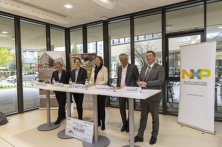 Five people, four men and one woman, stand behind high tables and speak to an audience