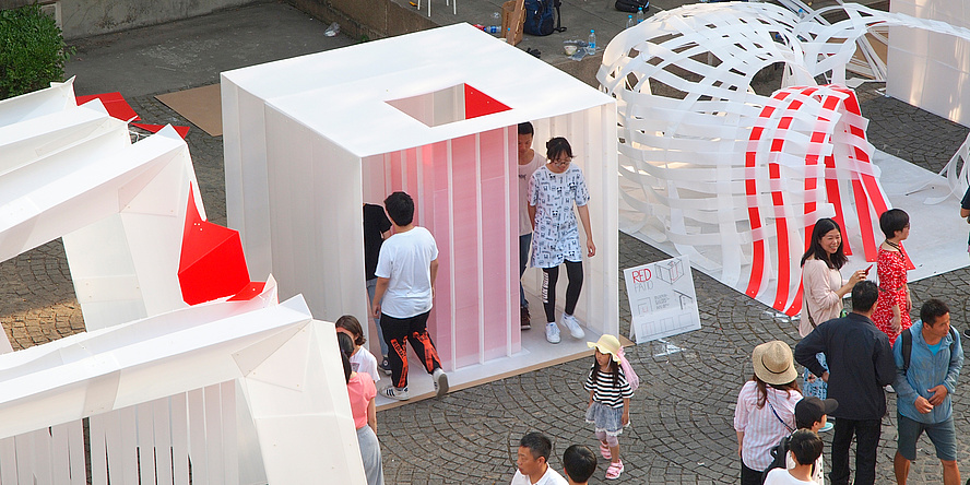 A small, white pavilion made of polypropylene hollow web sheets stands in the centre of the picture on a surface area of 2.25 by 2.25 metres, with a patio clad in red panels in the centre of the pavilion. Other pavilion variants are exhibited next to it. All around are Chinese visitors.