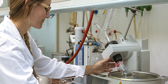 Eine Frau mit blonden Haaren, einer Schutzbrille und einem weißen Labormantel steht in einem Labor. Sie bedient ein Laborgerät, dass wie ein silberner Topf aussieht.
