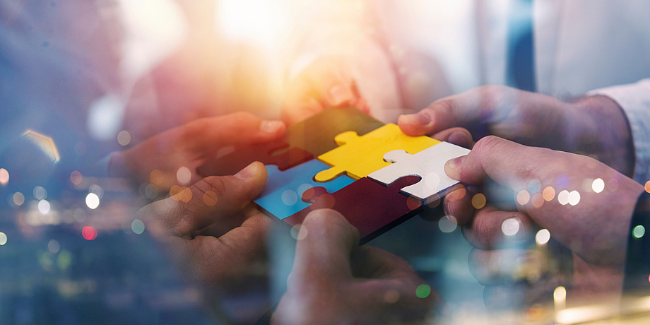 Four hands each hold a coloured puzzle piece. The hands try to put the puzzle pieces together.