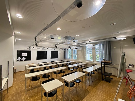 Blick in ein Klassenzimmer, unter der Decke sind Rohre und kreisrunde, transparente tellerartige Objekte zu sehen.