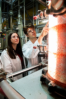Two people in lab coats are standing next to a large glass container filled with a liquid in which particles are floating.