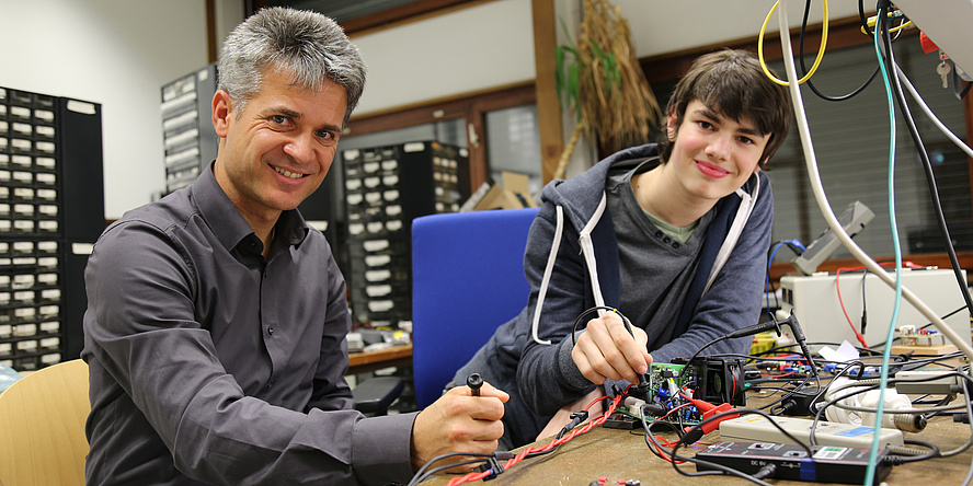 Ein älterer Mann und ein junger Mann sitzen an einem Tisch. Vor ihnen sind viele elektrische Geräte und Kabel zu sehen.