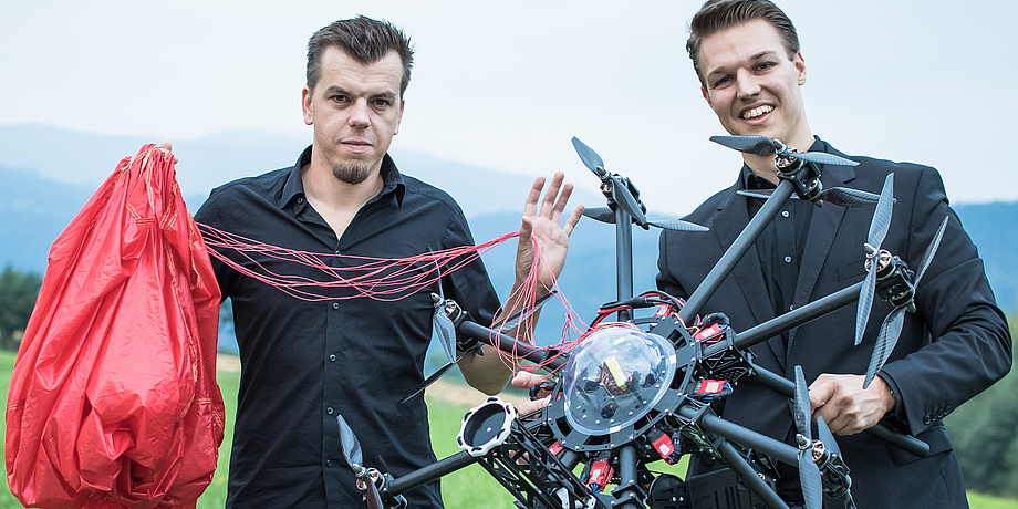Zwei junge Männer präsentieren ihre Erfindung, einen Rettungsfallschrim für Drohnen. Der Mann rechts im Bild hält eine schwarze Drohne, der Herr links im Bild einen roten Rettungsfallschirm.