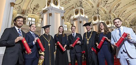 Acht Personen in festlicher Kleidung blicken in die Kamera und lächeln. Sechs von ihnen halten rote Rollen in der Hand.
