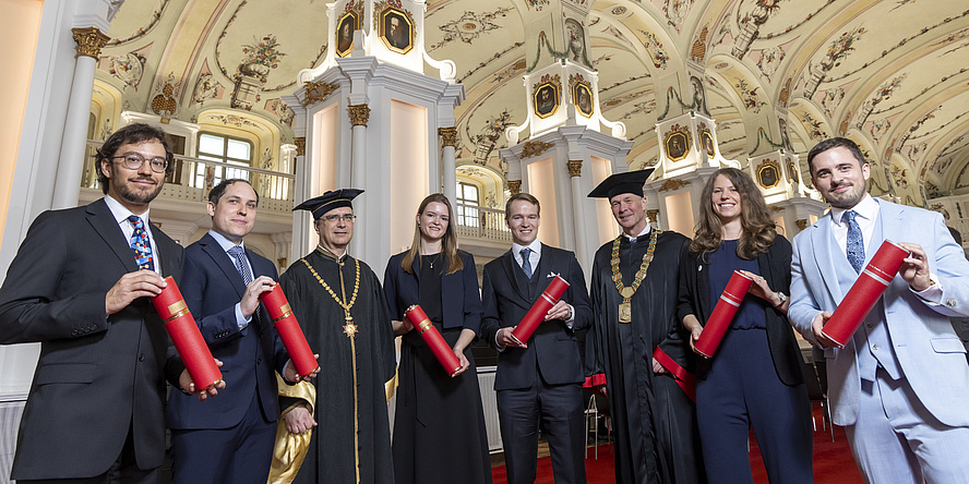 Acht Personen in festlicher Kleidung blicken in die Kamera und lächeln. Sechs von ihnen halten rote Rollen in der Hand.