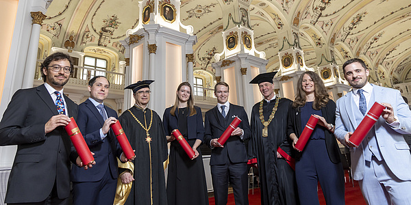 Acht Personen in festlicher Kleidung blicken in die Kamera und lächeln. Sechs von ihnen halten rote Rollen in der Hand.